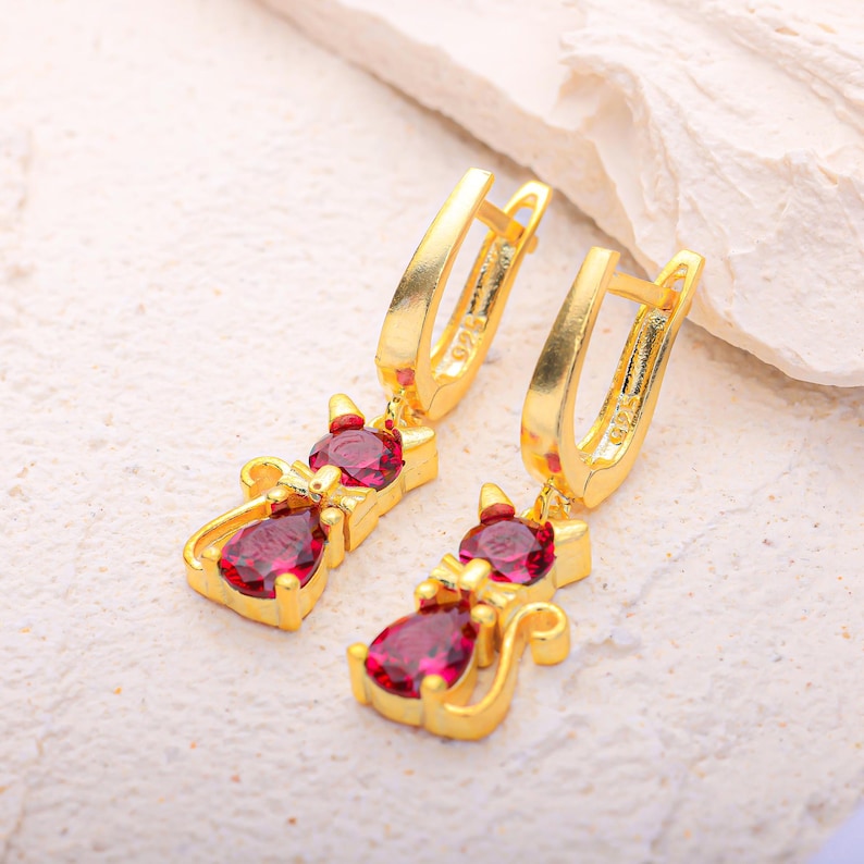 May include: A pair of gold-colored cat-shaped earrings with red gemstone accents. The earrings feature a lever back closure and are designed with a cat silhouette, including a head, body, and tail. The gemstones are a deep red color.