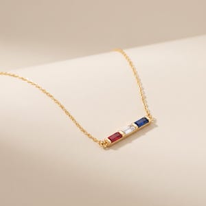 May include: A gold chain necklace featuring a horizontal bar pendant set with three rectangular baguette-cut gemstones in a row. The stones are arranged in a sequence of red, clear, and blue, held securely within a polished gold metal setting.