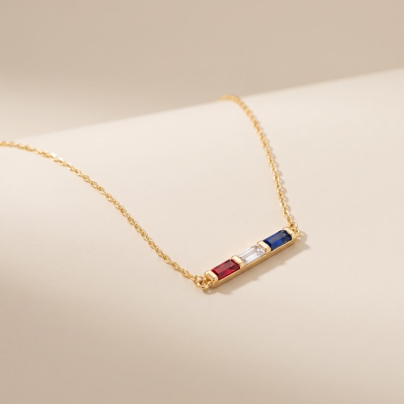 May include: A gold chain necklace featuring a horizontal bar pendant set with three rectangular baguette-cut gemstones in a row. The stones are arranged in a sequence of red, clear, and blue, held securely within a polished gold metal setting.