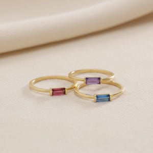 May include: Three gold rings with rectangular gemstones in pink, purple, and blue. The rings are set on a neutral-colored surface, with a soft, blurred background. The gemstones are set horizontally.