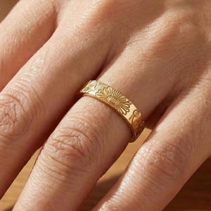 May include: A gold ring featuring a floral pattern with a sunburst motif. The ring is displayed on a finger, highlighting the intricate engravings. The ring's band is approximately 0.64 cm wide.