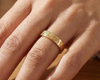 Peut inclure: Une bague dorée ornée d'un motif floral, notamment un motif de soleil. La bague est portée au doigt, mettant en valeur les gravures détaillées. La bande de la bague mesure environ 0,64 cm de large.