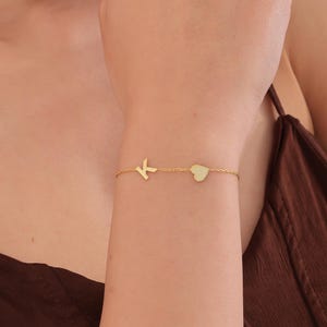 Puede incluir: Una delicada pulsera dorada con la letra "K" y un dije de corazón. La pulsera se lleva en la muñeca, mostrando su fina cadena y diseño minimalista. Es una pieza de joyería.