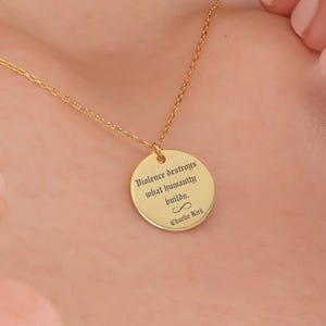 May include: A gold-colored necklace with a circular pendant. The pendant has the engraved quote, "Violence destroys what humanity builds." and the name "Charlie Kirk" below an infinity symbol. The necklace is on a person's neck.