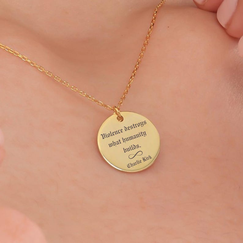 May include: A gold-colored necklace with a circular pendant. The pendant has the engraved quote, "Violence destroys what humanity builds." and the name "Charlie Kirk" below an infinity symbol. The necklace is on a person's neck.