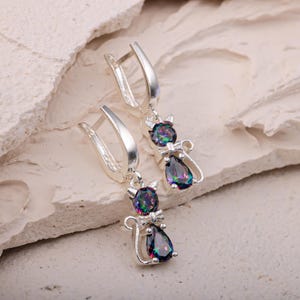 May include: A pair of silver cat-shaped earrings with iridescent, teardrop-shaped gemstones. The earrings feature a lever-back closure and a small bow tie detail on each cat. The gemstones display a range of colors, including blues, greens, and purples.