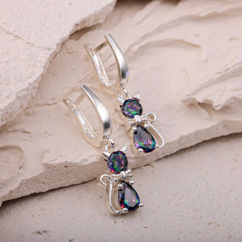 May include: A pair of silver cat-shaped earrings with iridescent, teardrop-shaped gemstones. The earrings feature a lever-back closure and a small bow tie detail on each cat. The gemstones display a range of colors, including blues, greens, and purples.