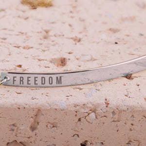 May include: A silver-colored bracelet with the word "FREEDOM" engraved on it. The bracelet is curved and appears to be made of metal. The background is a textured, light brown surface.