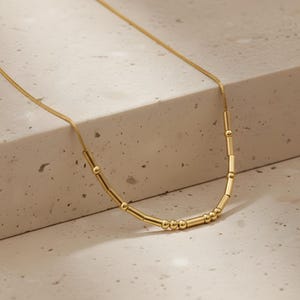 Op de afbeelding: Een goudkleurige ketting met een slangenketting en een reeks cilindrische en bolvormige kralen. De ketting wordt gepresenteerd op een neutrale ondergrond, waardoor de metallic glans en het minimalistische ontwerp worden benadrukt. De kralen zijn gelijkmatig verdeeld.