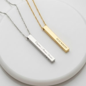 May include: Two personalized bar necklaces, one silver and one gold, displayed on a white surface. The silver necklace is engraved with "Together, Always, Forever." The gold necklace features Roman numerals and a name.