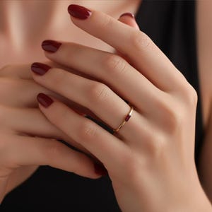 May include: A gold ring with a rectangular red gemstone. The ring is simple and elegant, with a thin band. The gemstone is set horizontally in the center of the ring. The ring is on a hand with red nail polish.