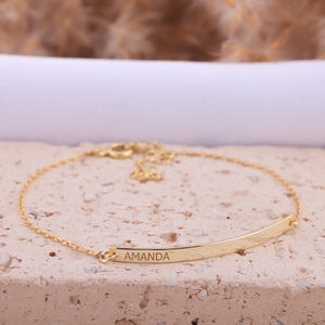 Op de afbeelding: Een gouden kettingarmband met een dunne, platte staafbedel gegraveerd met de naam "AMANDA".