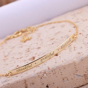 May include: A gold-colored necklace with a bar pendant. The pendant is inscribed with the words "thankful", "grateful", and "blessed". The necklace is displayed on a textured, light-colored surface.