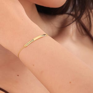 May include: A gold-colored bracelet with the word "FREEDOM" engraved on a rectangular bar. The delicate chain connects to the bar, creating a minimalist design. The bracelet is worn on a person's arm.