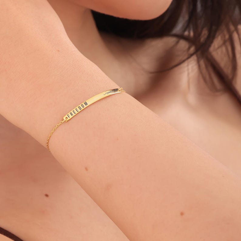 May include: A gold-colored bracelet with the word "FREEDOM" engraved on a rectangular bar. The delicate chain connects to the bar, creating a minimalist design. The bracelet is worn on a person's arm.