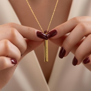 May include: A gold-colored vertical bar pendant necklace. The pendant has the Roman numerals IX.XX.MMXXIV engraved on it. The necklace is held by a person with dark red nail polish.