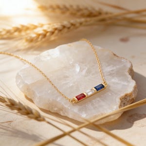 May include: A gold chain necklace featuring a horizontal bar pendant set with three rectangular baguette-cut gemstones in a row, specifically a red stone, a clear white stone, and a deep blue stone, attached to a delicate gold link chain.
