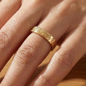 Puede incluir: Un anillo dorado con un diseño floral. El anillo presenta un patrón repetitivo de flores en forma de sol y otros elementos decorativos. El anillo se lleva en un dedo, mostrando los detalles grabados. El diseño es intrincado.