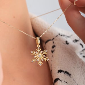 May include: A gold-colored snowflake pendant necklace. The pendant is adorned with small, clear gemstones and hangs from a delicate chain. The necklace is being held up, showcasing the intricate design and sparkle of the jewelry.