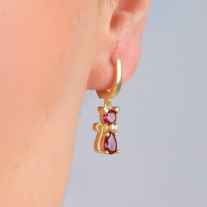 May include: Gold-colored cat-shaped earrings with red gemstones. The earrings feature a small cat design with a round gemstone at the head and a teardrop-shaped gemstone at the bottom. The earrings are attached to a gold-colored hoop.