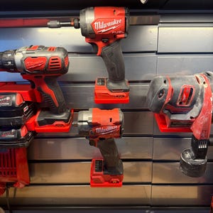 May include: Four red and black power tools are mounted on a silver wall. The tools are all Milwaukee brand and include a drill, an impact driver, a wrench, and a sander. The tools are all hanging on a wall-mounted tool organizer.