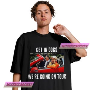 Może przedstawiać: Czarna koszulka z białym napisem "GET IN DOGS WE'RE GOING ON TOUR". Koszulka przedstawia grafikę trzech os&oacute;b w czerwonym kabriolecie. Koszulka ma okrągły dekolt.