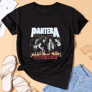 Op de afbeelding: Zwart T-shirt met de bandnaam "PANTERA" in wit boven een bandafbeelding. Daaronder staat de tekst "COWBOYS FROM HELL" in een rode doos. Het shirt wordt gecombineerd met witte sandalen en een blauwe spijkerbroek.