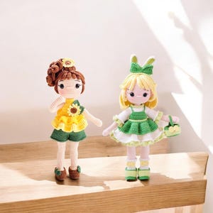Puede incluir: Dos muñecas de crochet, una con cabello castaño y un vestido amarillo de girasol, la otra con cabello rubio y un vestido verde con encaje blanco.