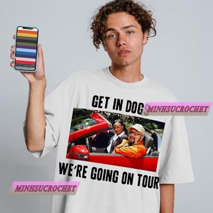 Może przedstawiać: Biała koszulka z grafiką czerwonego kabrioletu z dwiema osobami w środku. Tekst "GET IN DOG WE'RE GOING ON TOUR" jest wydrukowany na czarno. Osoba trzyma smartfon z kolorowym wzorem.