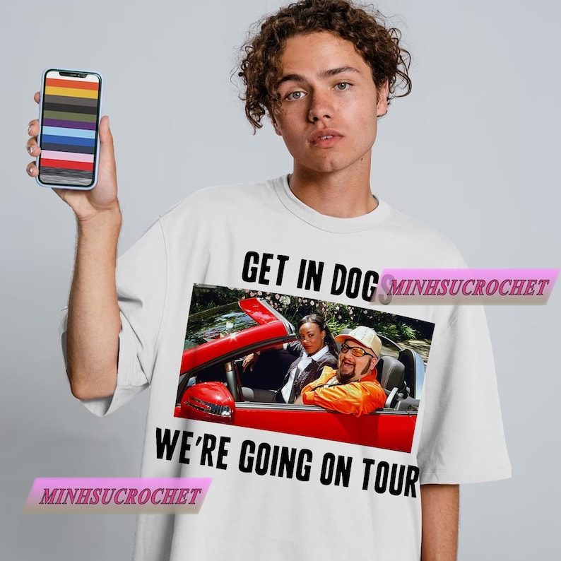 Może przedstawiać: Biała koszulka z grafiką czerwonego kabrioletu z dwiema osobami w środku. Tekst "GET IN DOG WE'RE GOING ON TOUR" jest wydrukowany na czarno. Osoba trzyma smartfon z kolorowym wzorem.