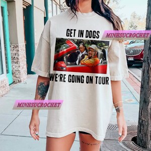 Może przedstawiać: Kremowy t-shirt z grafiką czerwonego kabrioletu z dwiema osobami. Tekst "GET IN DOGS" znajduje się nad grafiką, a "WE'RE GOING ON TOUR" poniżej. Koszulka ma luźny kr&oacute;j.