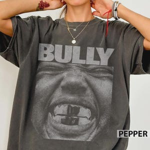 Op de afbeelding: Donkergrijs t-shirt met het woord "BULLY" in lichtgrijze letters op de borst. Daaronder een zwart-wit afbeelding van een gezicht. Het woord "PEPPER" staat rechtsonder. Casual stijl.