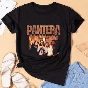 May include: Black t-shirt featuring the band Pantera. The shirt has a graphic of the band members with the band's name in a distressed, textured font. The shirt is paired with white sandals and denim shorts.