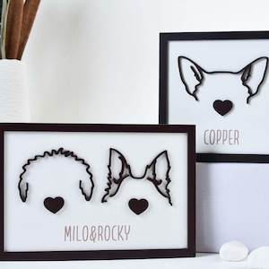 Op de afbeelding: Twee ingelijste hondenportretten met zwarte contouren van hondengezichten en hartaccenten. Eén lijst zegt "COPPER" en de andere "MILO & ROCKY". De lijsten zijn donkerbruin en staan op een wit oppervlak.