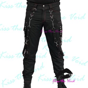May include: Black pants with red stitching, featuring multiple straps, chains, and metal accents. The pants have cargo pockets and decorative metal rings. The overall design has a punk or alternative aesthetic.