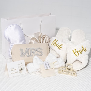 Può includere: Un sacchetto regalo bianco pieno di accessori da sposa. Include una mascherina per dormire, un elastico per capelli, un beauty case con "MRS" in strass, pantofole soffici con "Bride" in oro, una fascia per capelli, un biglietto con "Best Wishes" e una forcina per capelli con perle.