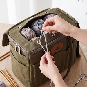 May include: An olive green canvas yarn storage bag featuring a zippered top, multiple exterior pockets, and metal grommets for feeding yarn through. The bag contains several balls of yarn in neutral tones and has a brown leather-like patch.