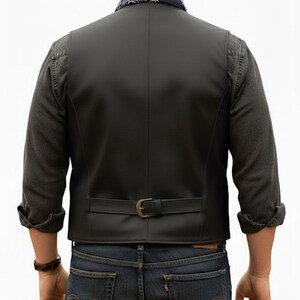 Puede incluir: Un chaleco negro de acabado liso con una tira trasera horizontal y hebilla met&aacute;lica. El chaleco se lleva sobre una camisa de manga larga gris oscuro con pu&ntilde;os vueltos, combinado con vaqueros azul oscuro y un pa&ntilde;uelo azul con motivos estampados.