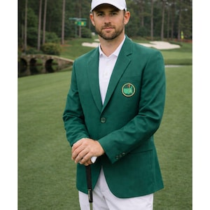 Puede incluir: Una persona con una chaqueta verde con un emblema circular, camisa blanca y gorra blanca, sosteniendo un palo de golf en un campo verde. La chaqueta tiene cierre de dos botones y solapa.