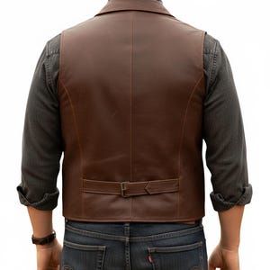 Puede incluir: Un chaleco de cuero marr&oacute;n con cuello de muesca, acabado liso y una correa ajustable con hebilla de metal en la parte inferior de la espalda. El chaleco se lleva sobre una camisa de manga larga gris oscuro y vaqueros azul oscuro, dise&ntilde;o cl&aacute;sico.