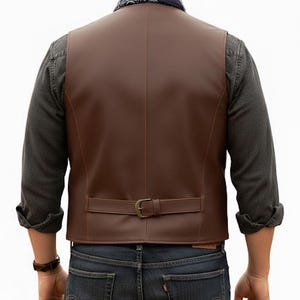 Puede incluir: Un chaleco de cuero marr&oacute;n con acabado liso, una costura vertical en el centro de la espalda y una correa ajustable en la cintura con hebilla met&aacute;lica. El chaleco se lleva sobre una camisa de manga larga gris oscuro y vaqueros azul oscuro.