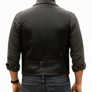 Puede incluir: Un chaleco de cuero negro con una correa de ajuste trasera con hebilla de metal, usado sobre una camisa de manga larga gris oscuro. El chaleco tiene un acabado liso y un corte entallado, combinado con pantalones vaqueros azul oscuro y un pa&ntilde;uelo.