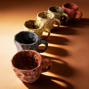 以下が含まれることがあります： 赤、緑、青、黄色など、さまざまな色の手作りのセラミックマグカップ6個セット。各マグカップはユニークな質感の表面と丸みを帯びたハンドルを備えています。マグカップは斜めに配置されており、それぞれのデザインが際立っています。