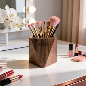 May include: A wooden makeup brush holder with a geometric design, holding various makeup brushes with pink and gold accents. The holder is engraved with the name "Amelia". Lipsticks and other makeup items are also visible.