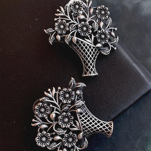 Puede incluir: Dos broches plateados, cada uno con forma de cesta rebosante de flores y follaje. Los broches tienen una estética vintage detallada, con un fondo oscuro. El diseño de la cesta presenta un patrón de enrejado.