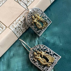 May include: Silver-coloured earrings with a gold-coloured figure design. The earrings have a detailed, openwork frame with a stylized figure in the centre. The figures appear to be dancing or in a celebratory pose. The earrings are displayed on a patterned surface.