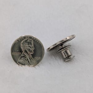 May include: A silver-colored lapel pin with a penny design. The pin features a detailed image of Abraham Lincoln and the words "In God We Trust" and "Liberty". The pin is shown next to a penny.