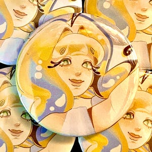 May include: A collection of round, glossy buttons featuring a smiling cartoon character with flowing blonde hair, green eyes, and a light blue and yellow color scheme. The buttons have a pin back.