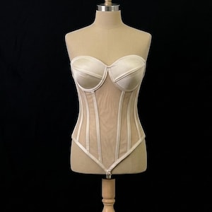 Può includere: Un corsetto beige senza spalline con scollatura a cuore e dettagli di stecche verticali. Il corpetto è realizzato in un materiale simile al raso, con pannelli in rete trasparente e stecche verticali. Il corsetto è esposto su un manichino.
