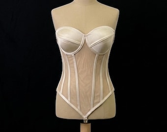 May include: A beige strapless corset with a sweetheart neckline and vertical boning details. The bodice is made of a satin-like material, with sheer mesh panels and vertical boning. The corset is displayed on a mannequin against a black background.